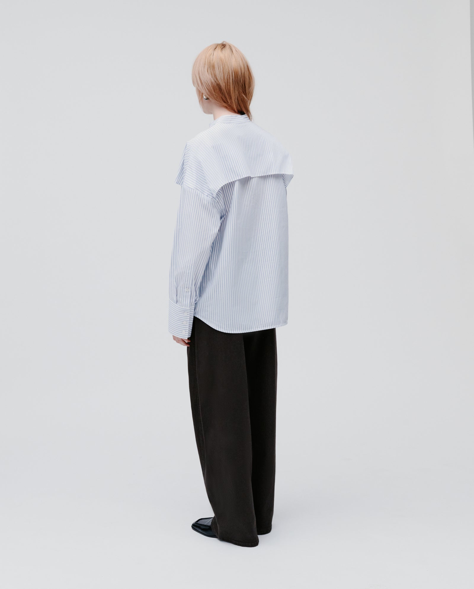A person with light hair, seen from behind, wears the IVY OAK EVE Blouse—an oversized striped shirt with buttons—paired with dark trousers.