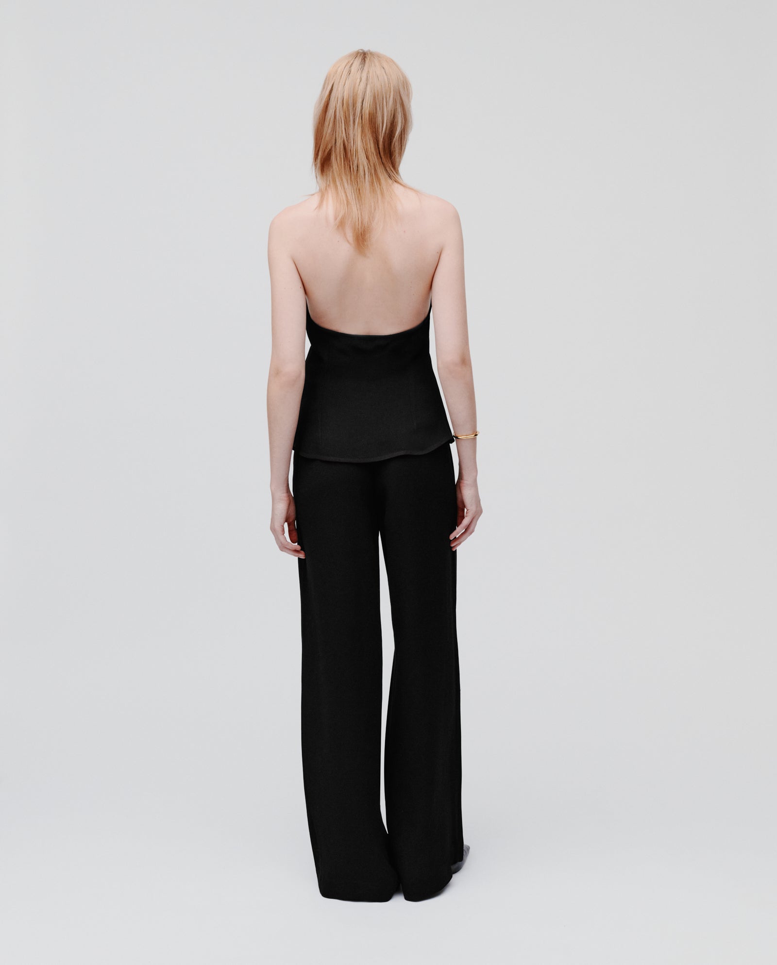 A blonde woman, seen from behind, wears the IVY OAK TANSY Top—a neckholder style in crepe fabric—paired with black pants.