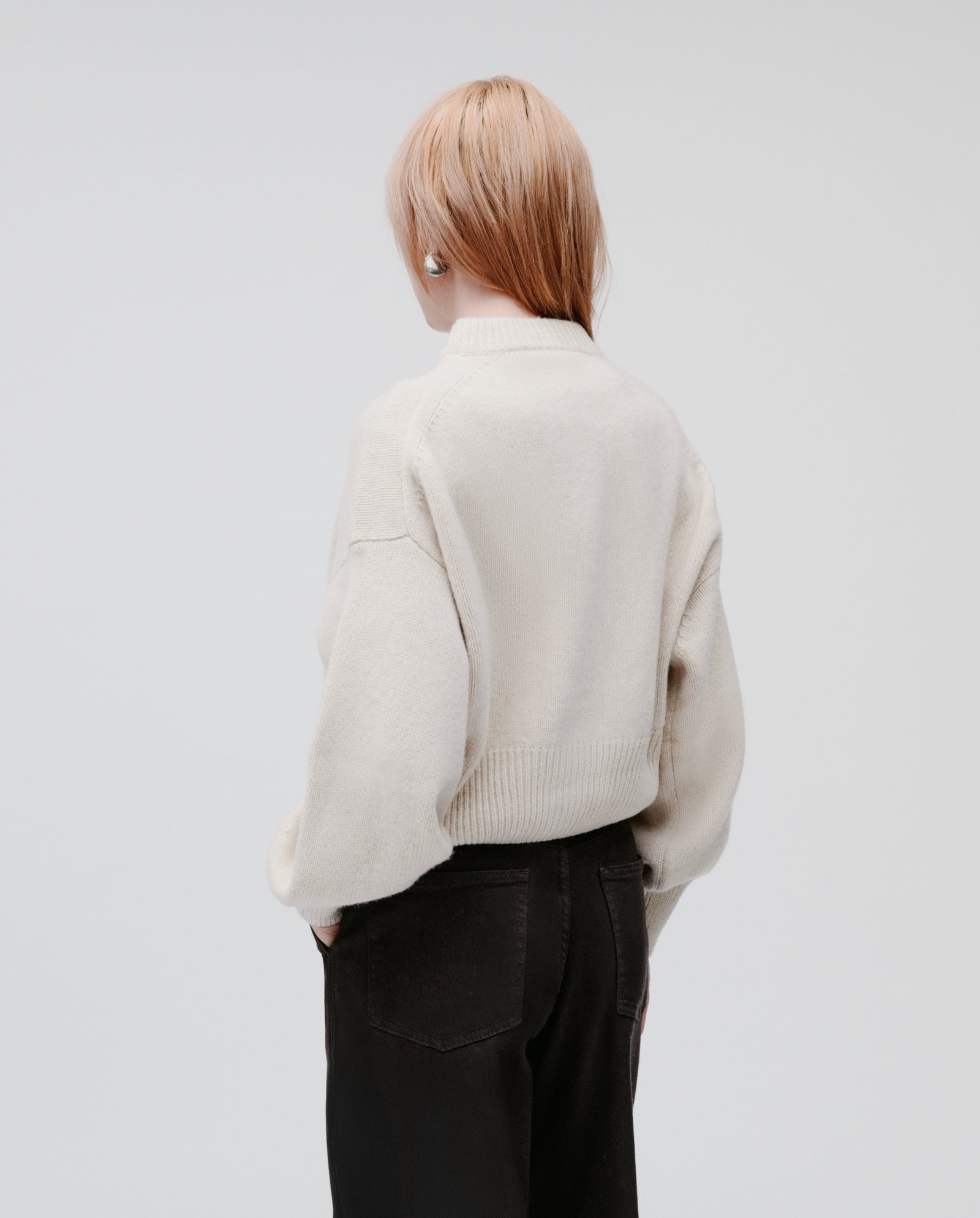 Person with light brown hair stands with their back to the camera, wearing the IVY OAK KARMA Cardigan—an oversized cream cashmere-blend piece with wide sleeves—paired with black pants.