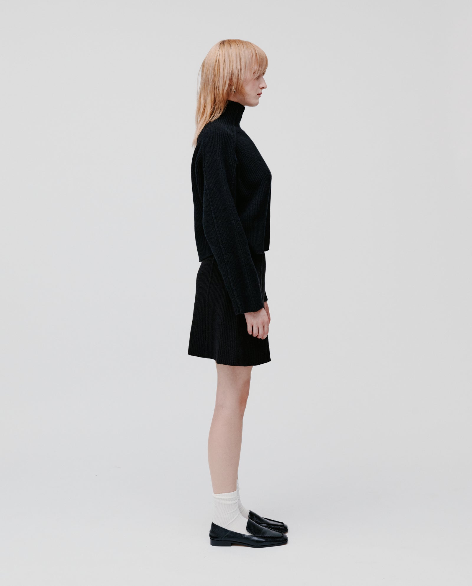 A person with light hair wears the IVY OAK KEEVA Jumper in an oversized fit, paired with a skirt and loafers, standing in profile against a plain background.