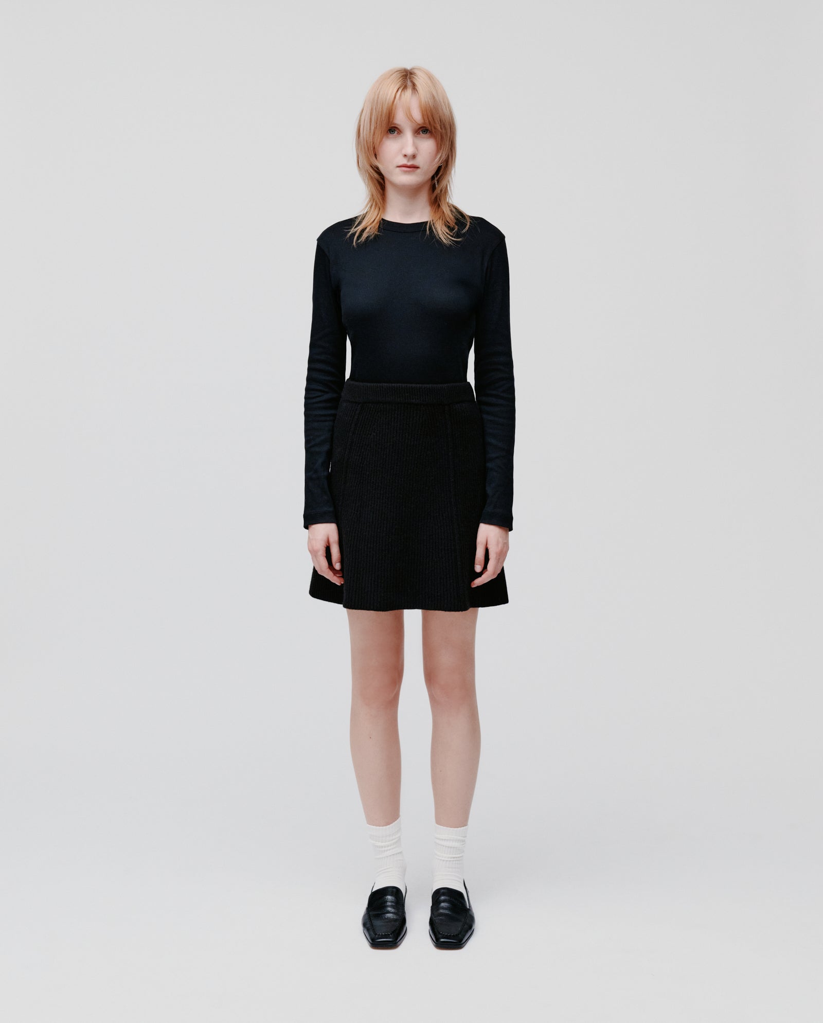 A woman faces forward wearing the IVY OAK KEENA Skirt with a black long-sleeve top, white socks, and black shoes.