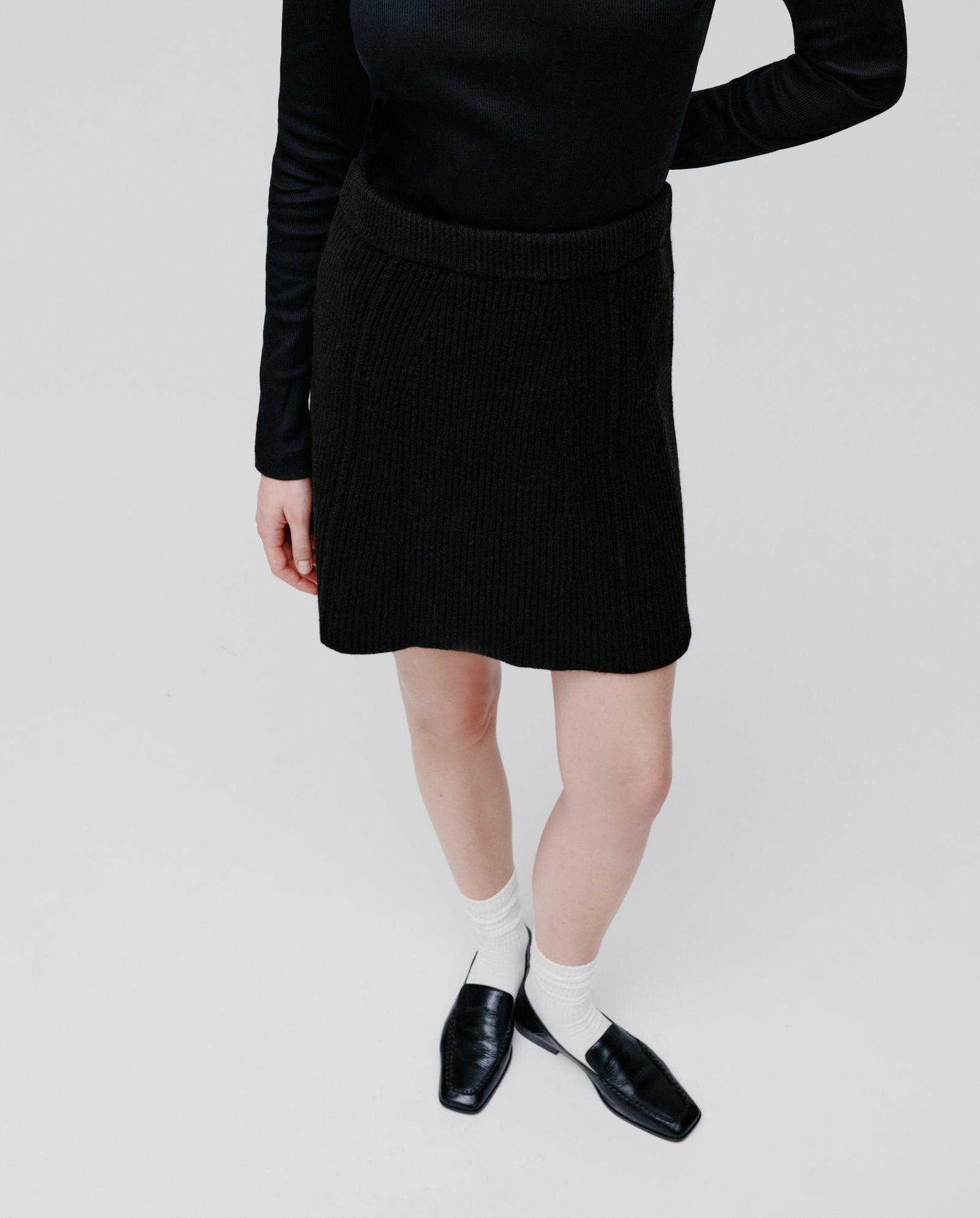A person wearing a black top, KEENA Skirt by IVY OAK, white socks, and black shoes stands against a plain background.