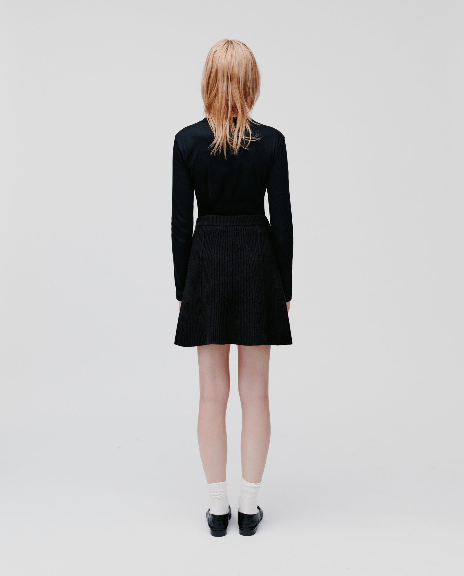 A blonde woman in a black outfit featuring the IVY OAK KEENA Skirt and white socks stands facing away from the camera.