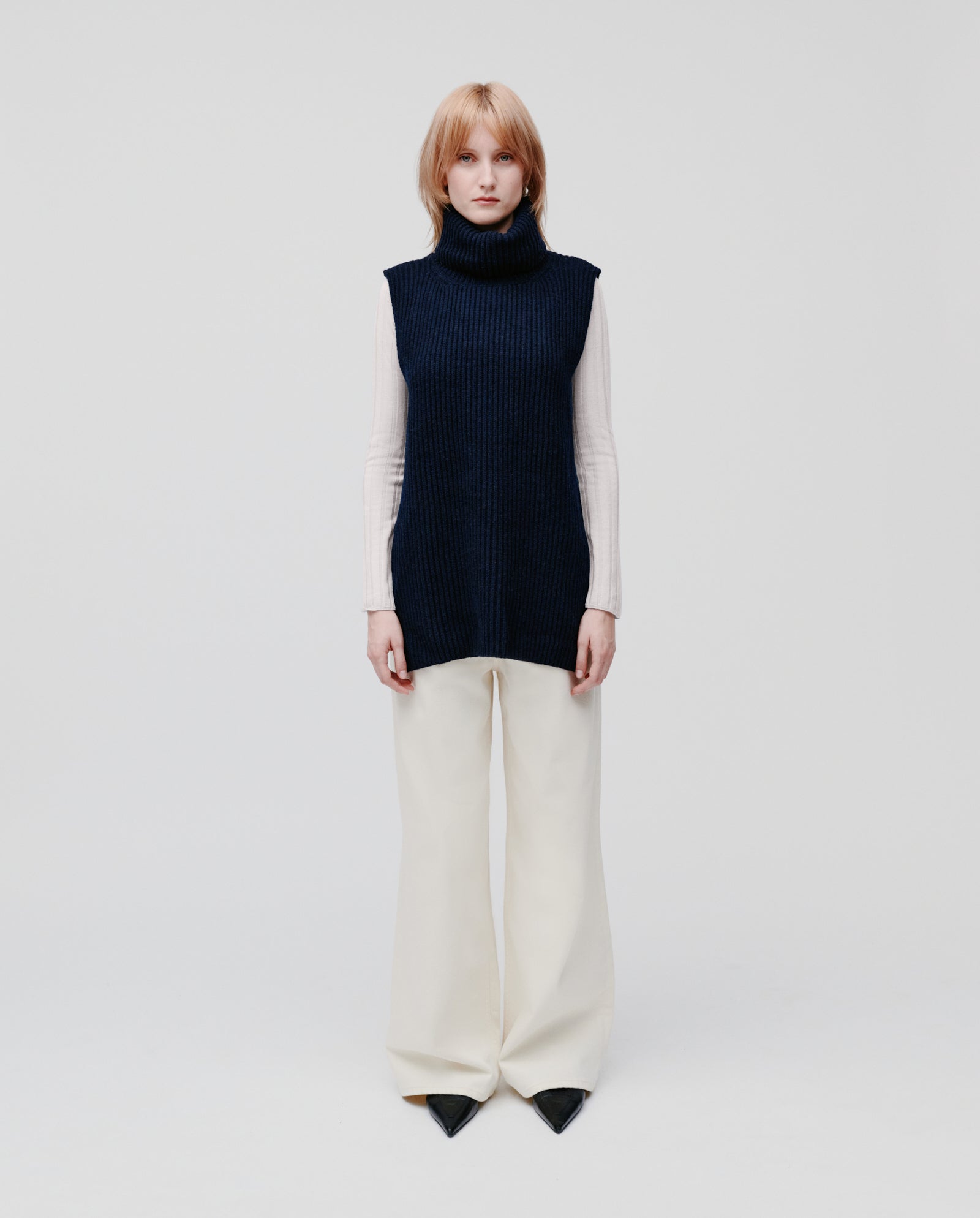 A woman with blonde hair wears the IVY OAK KYOMI Pullunder, a navy chunky wool high-neck vest, layered over a white top and wide-leg white pants.