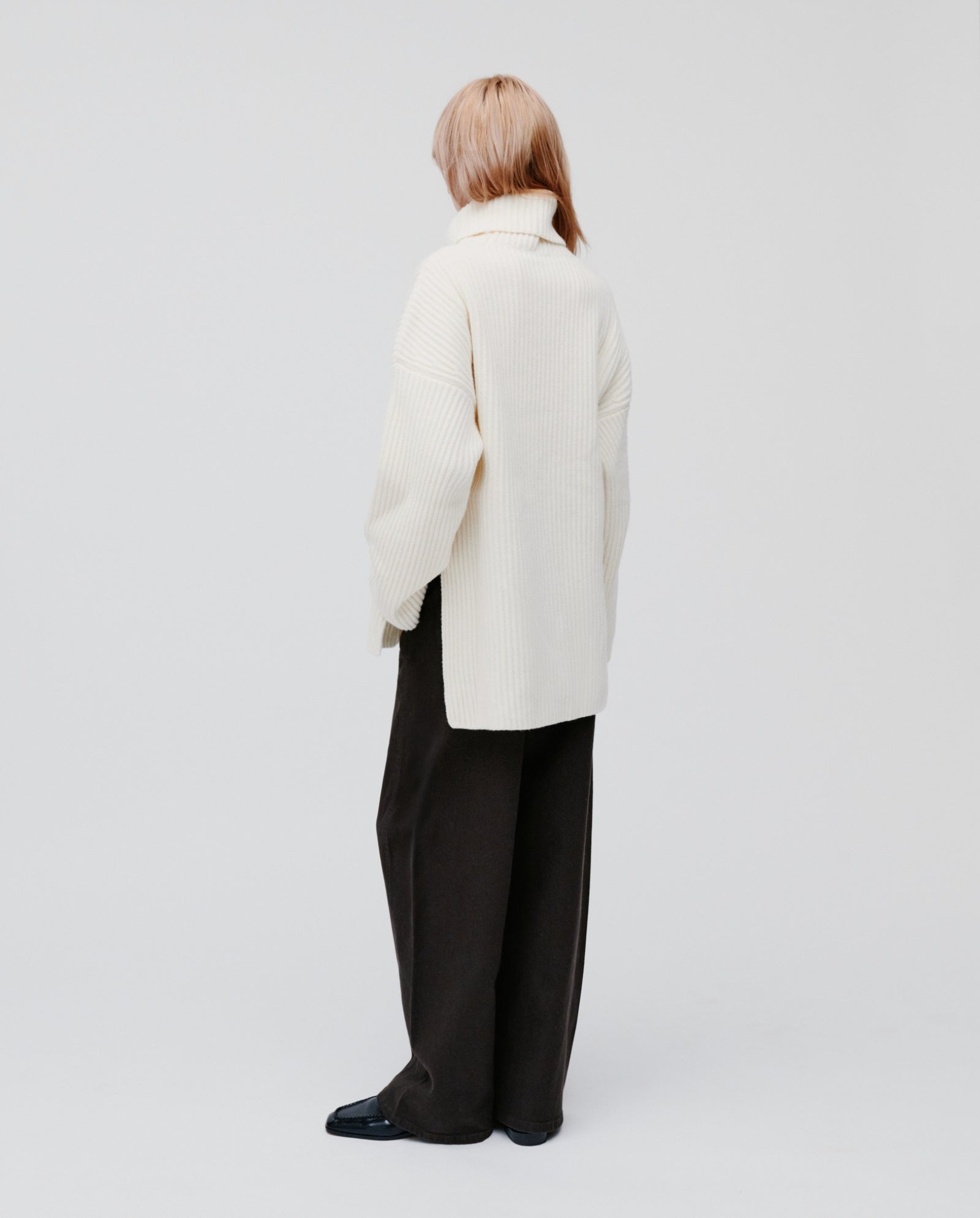 A person stands backwards against a plain background, wearing the IVY OAK KYOMI ANN chunky knitted jumper in cream with loose black pants.