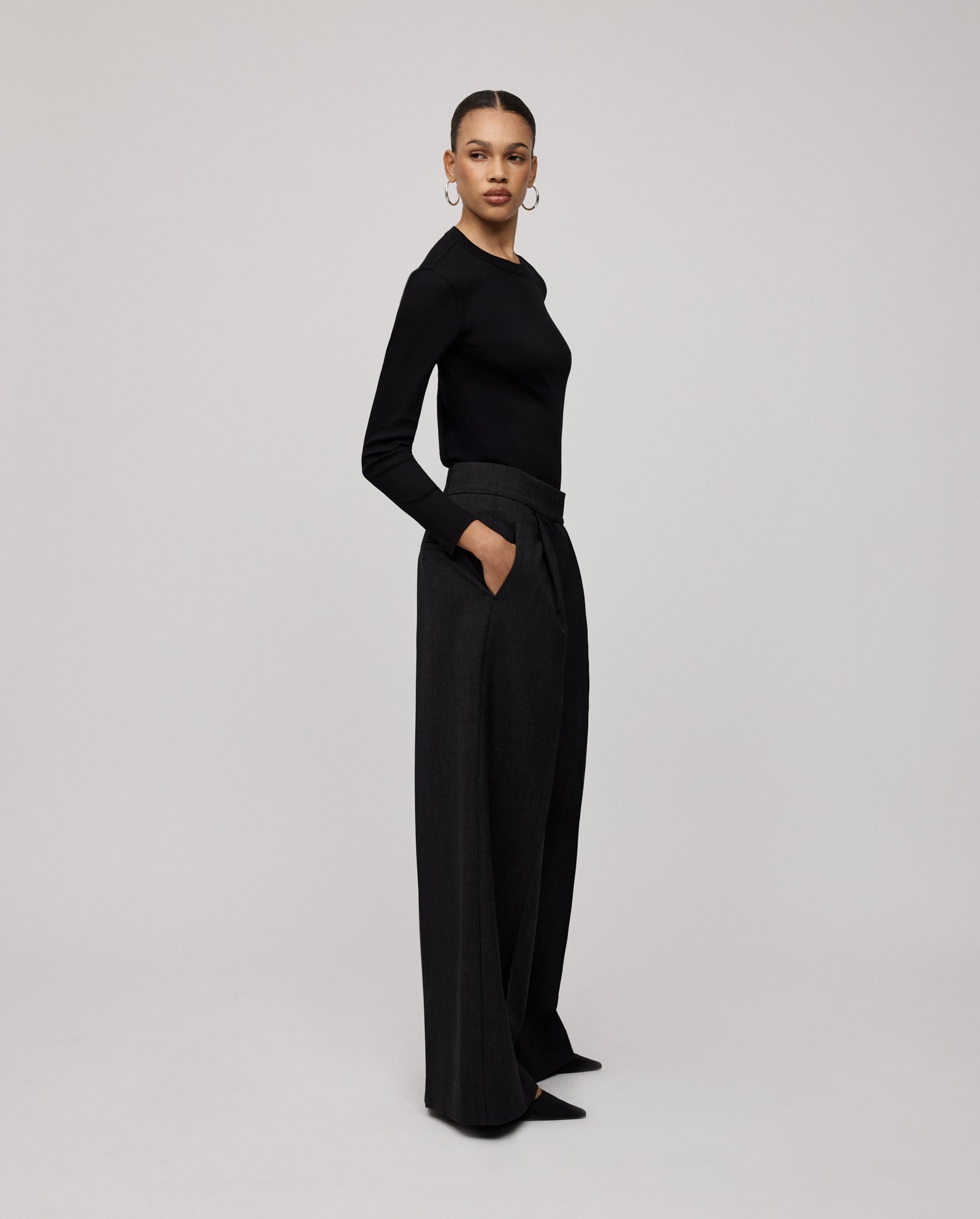 A woman stands with her hands in her pockets, wearing wide-leg PRESCILLIA SUE Pants by IVY OAK and a black long-sleeve top.