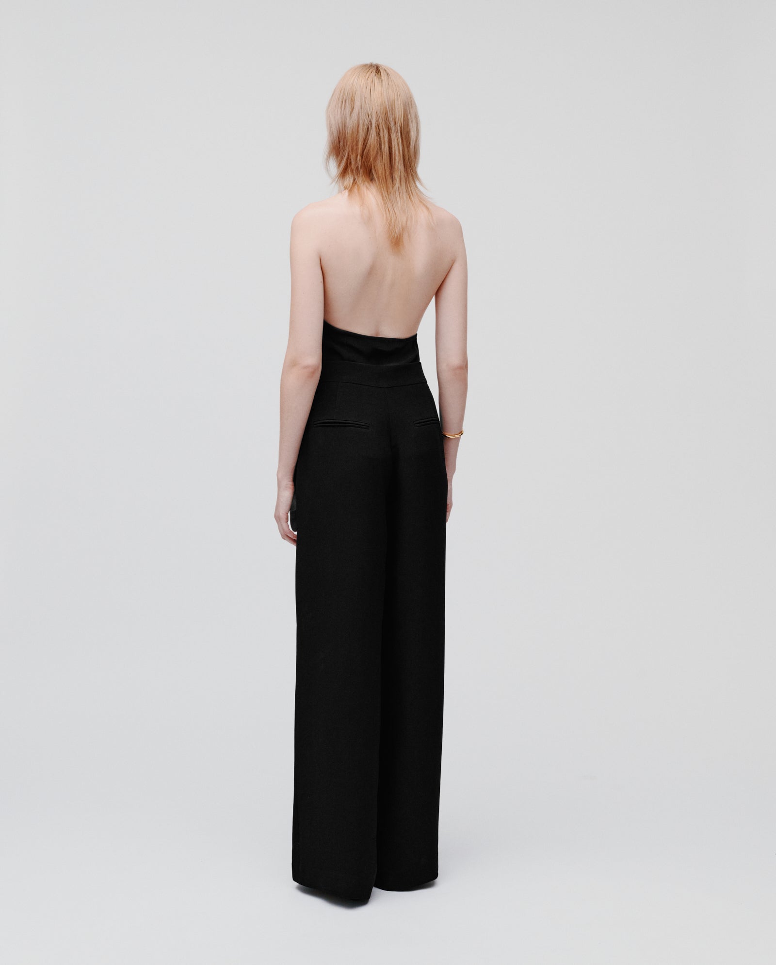 A person stands with their back to the camera, wearing IVY OAKs PHILINE SUE Pants—strapless, black, wide-leg trousers—set against a minimalistic light background.