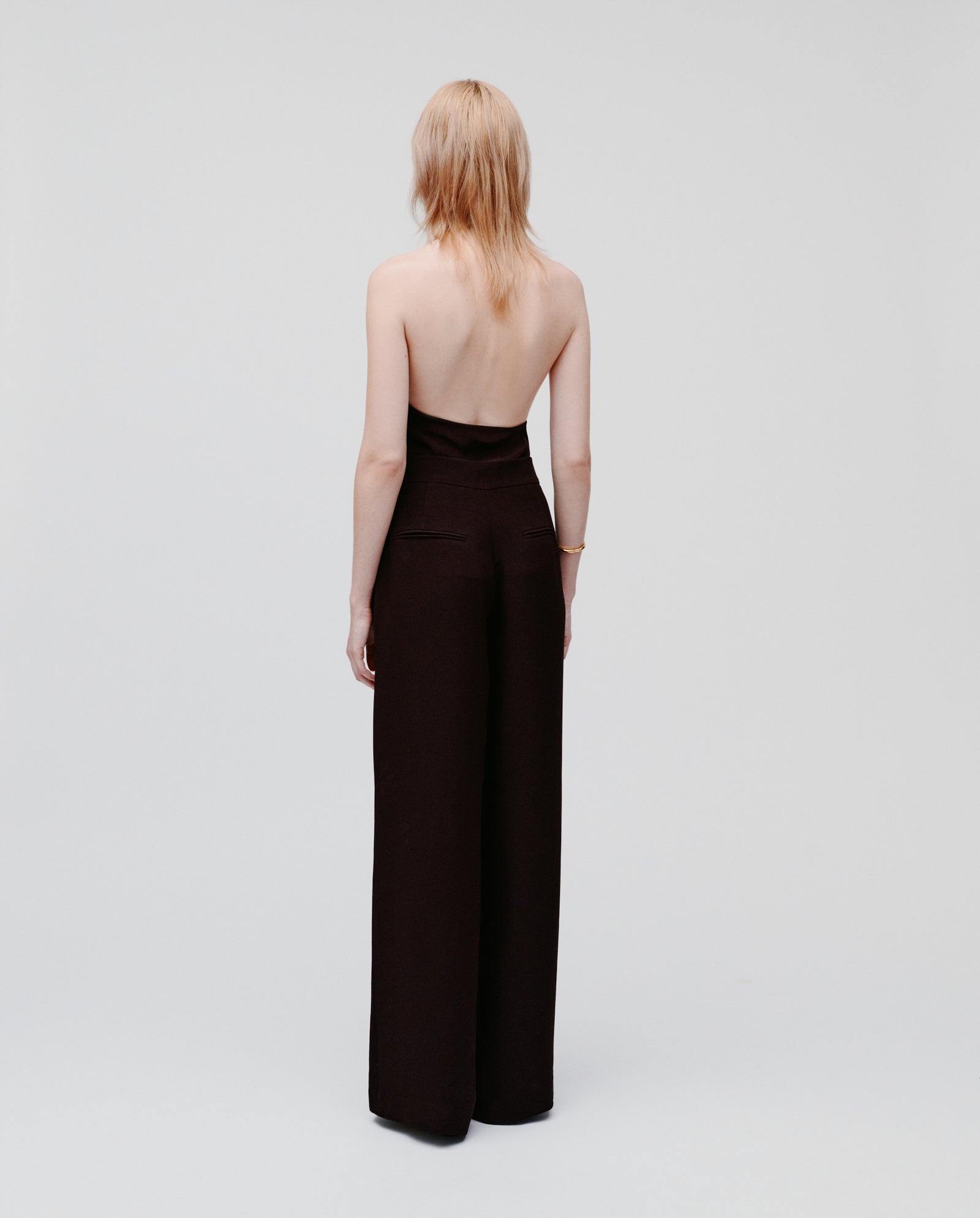 A person with light hair wears the IVY OAK PHILINE SUE Pants, a strapless black jumpsuit with wide-leg trousers, standing and facing away against a plain background.