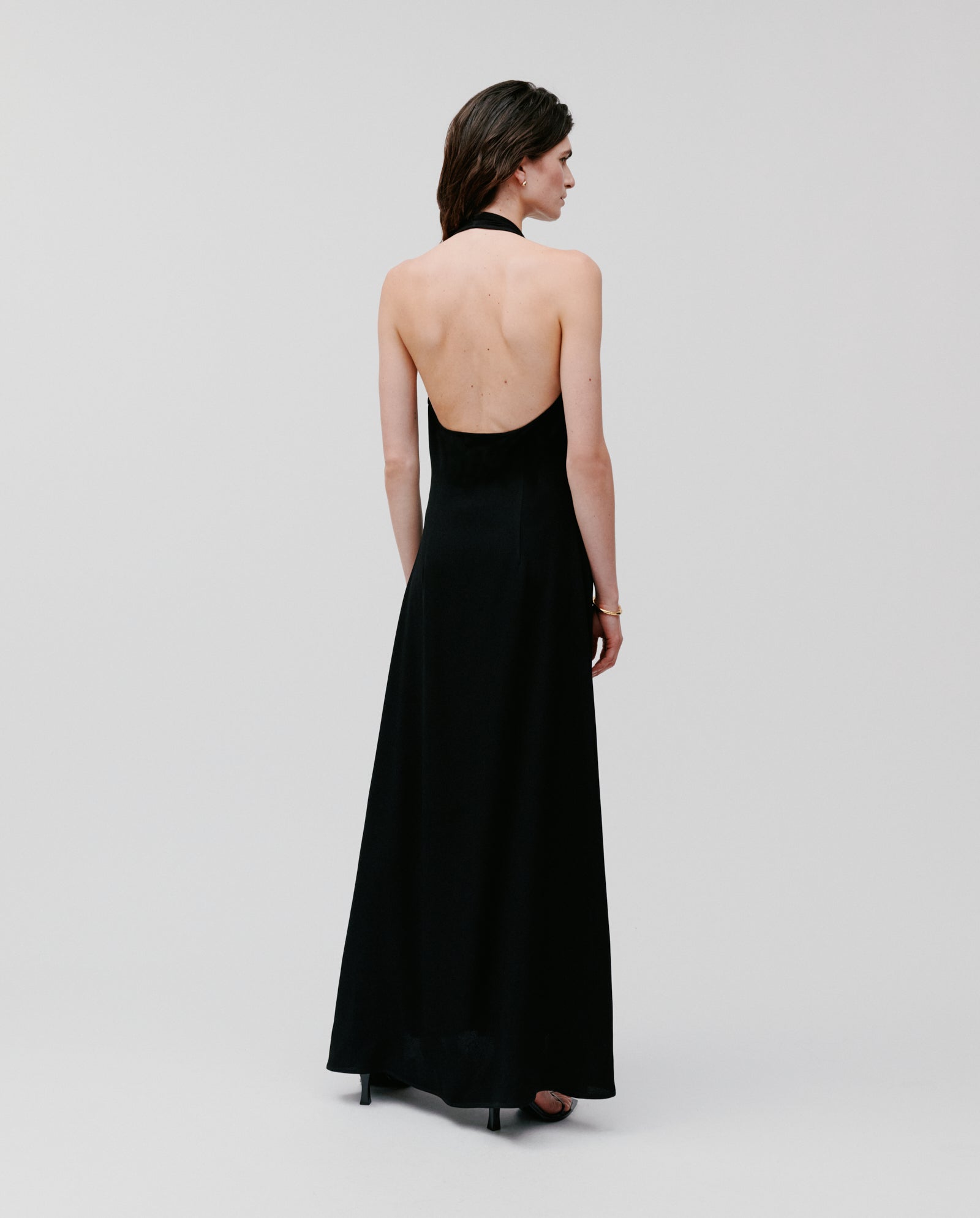 A woman stands facing away against a plain light background, wearing the IVY OAK NESTA Dress—a backless, black neckholder style crafted from smooth crepe fabric.