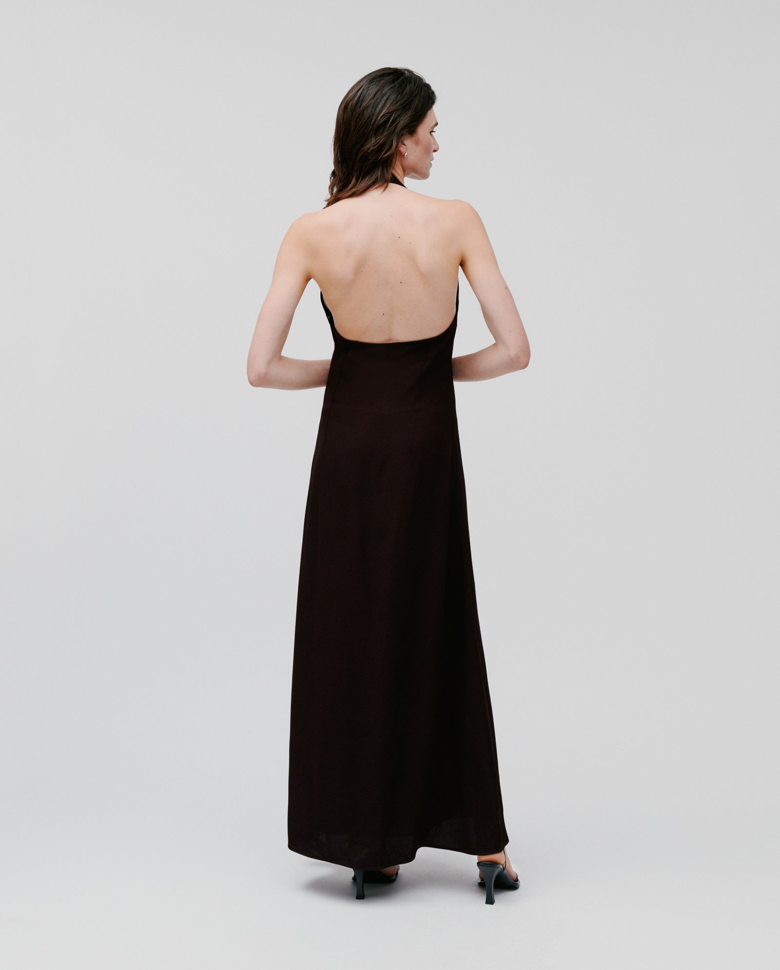 A woman stands with her back to the camera, wearing the IVY OAK NESTA Dress—a strapless, floor-length black crepe gown—paired with heels.