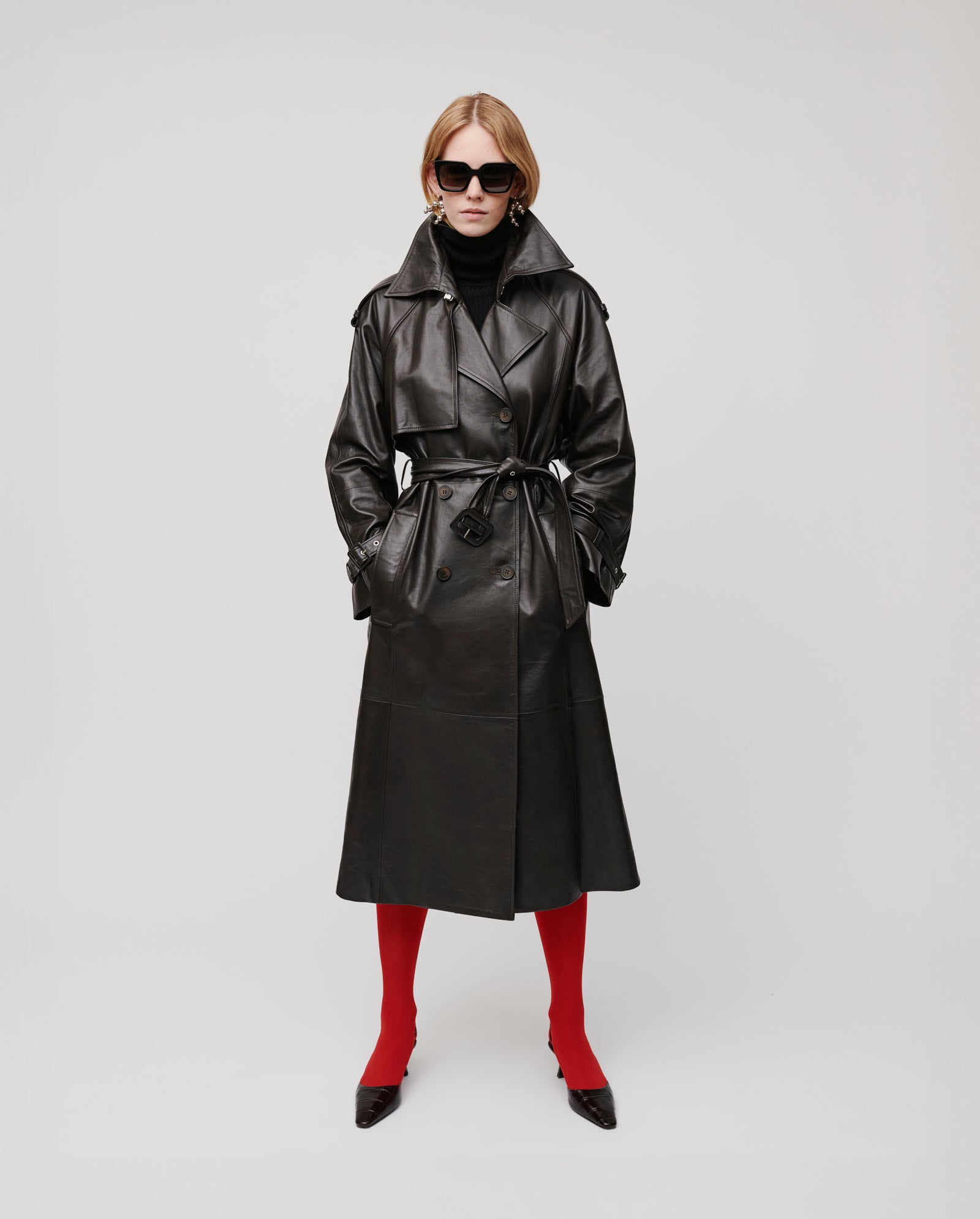 A woman in the IVY OAK LINDA black double-breasted leather trench coat, paired with a black turtleneck, red tights, black heels, and sunglasses.