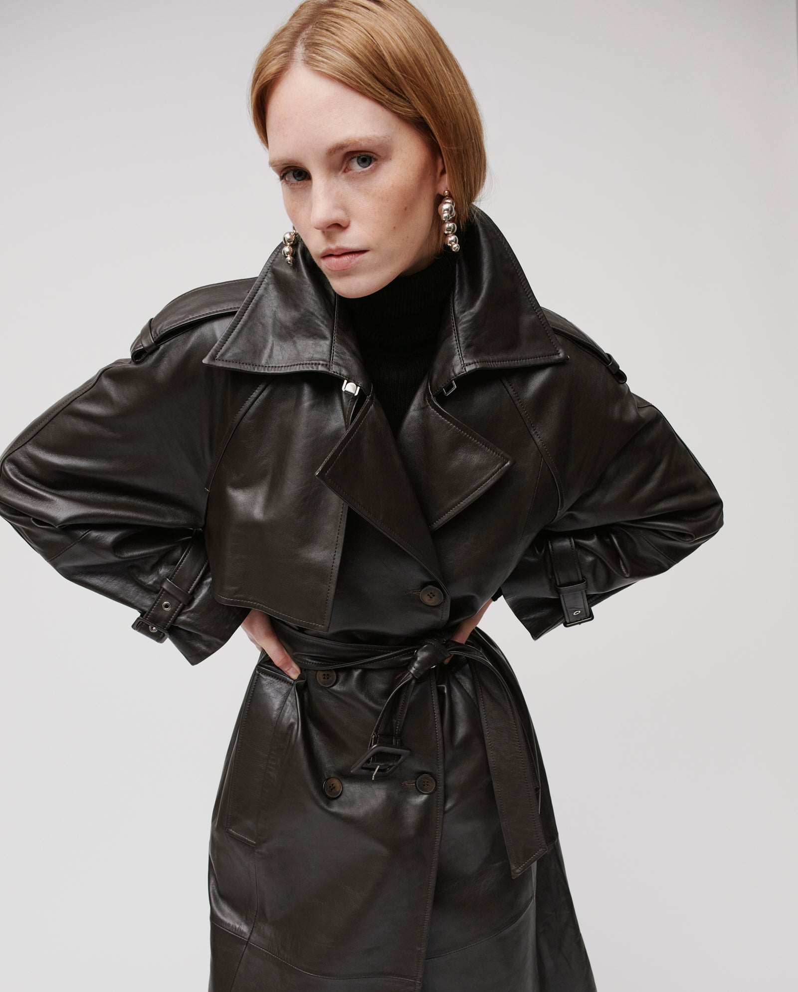 A woman stands confidently with hands on hips, modeling the oversized LINDA Coat by IVY OAK, looking directly at the camera.