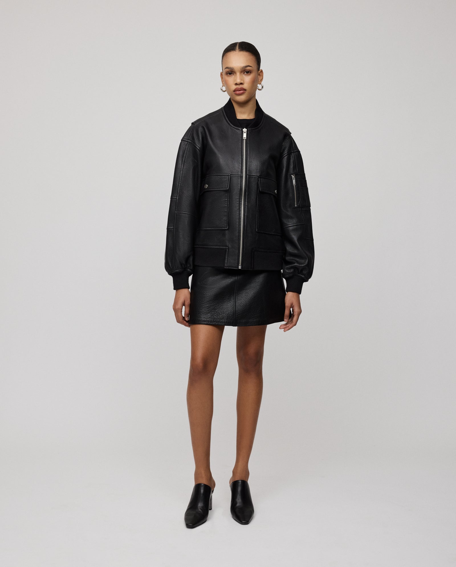 A woman wears the IVY OAK LARA Jacket, an oversized bomber crafted from premium hide, styled with a skirt and backless shoes, standing against a plain white background.