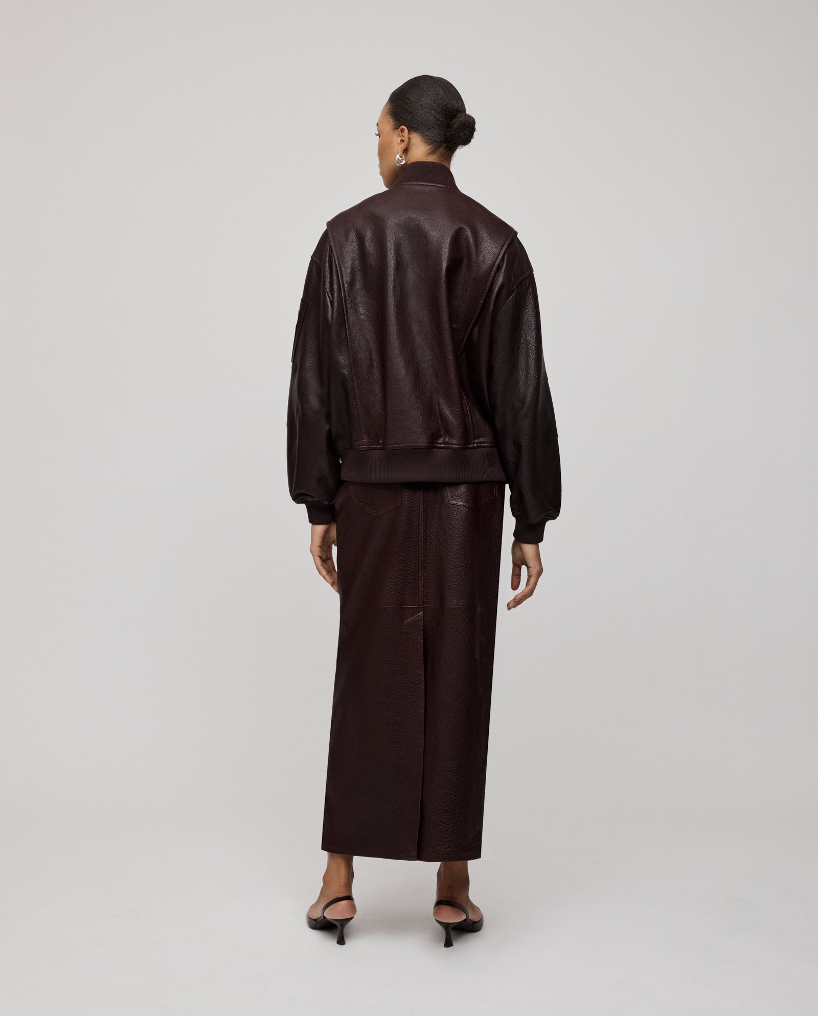 A woman stands facing away in a studio wearing the IVY OAK LARA Jacket, an oversized bomber with a patched sleeve pocket, paired with a dark brown premium hide skirt.