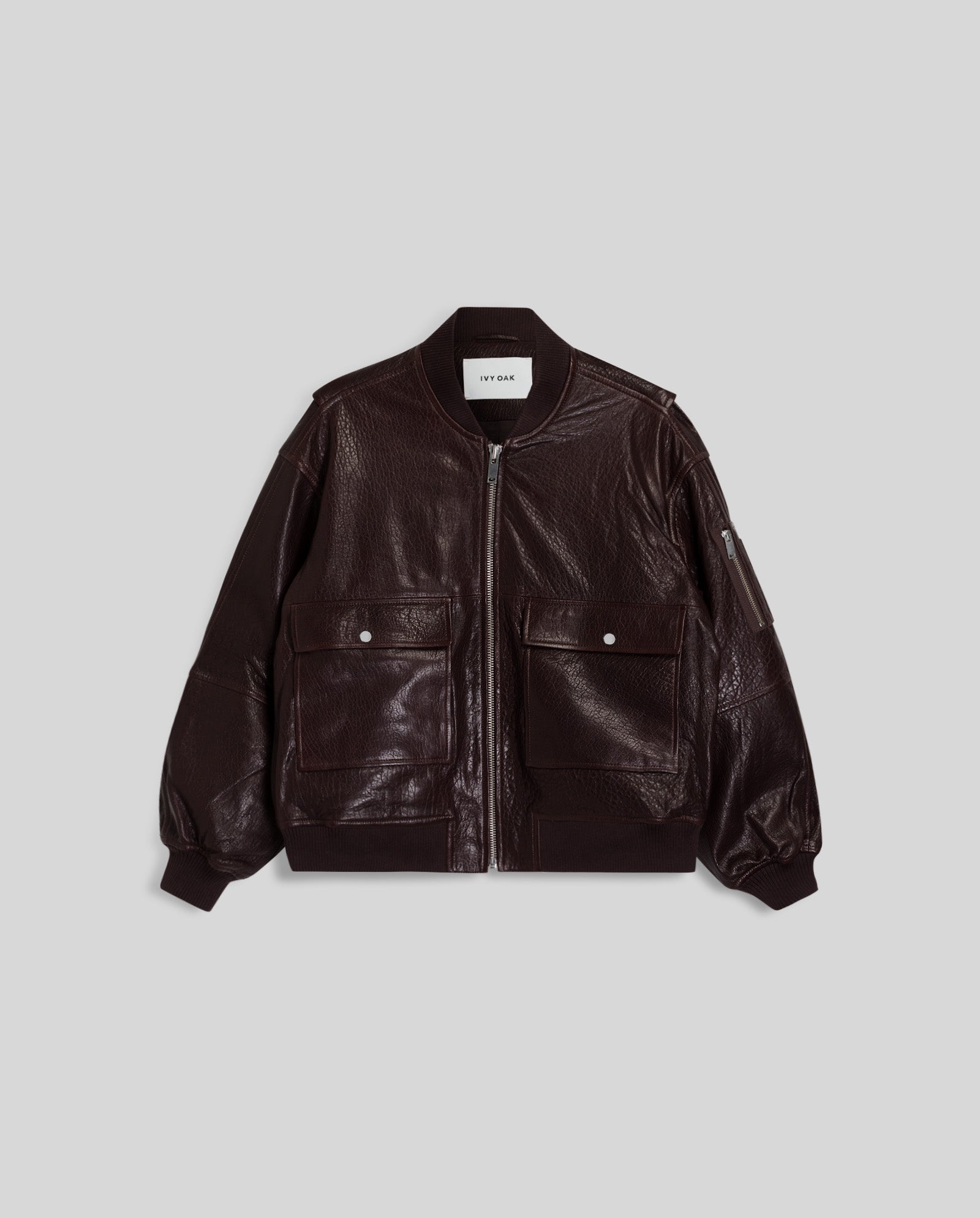 The LARA Jacket by IVY OAK is a dark brown oversized bomber crafted from premium hide, featuring a front zipper, large flap pockets, a patched sleeve pocket, and ribbed cuffs and hem.