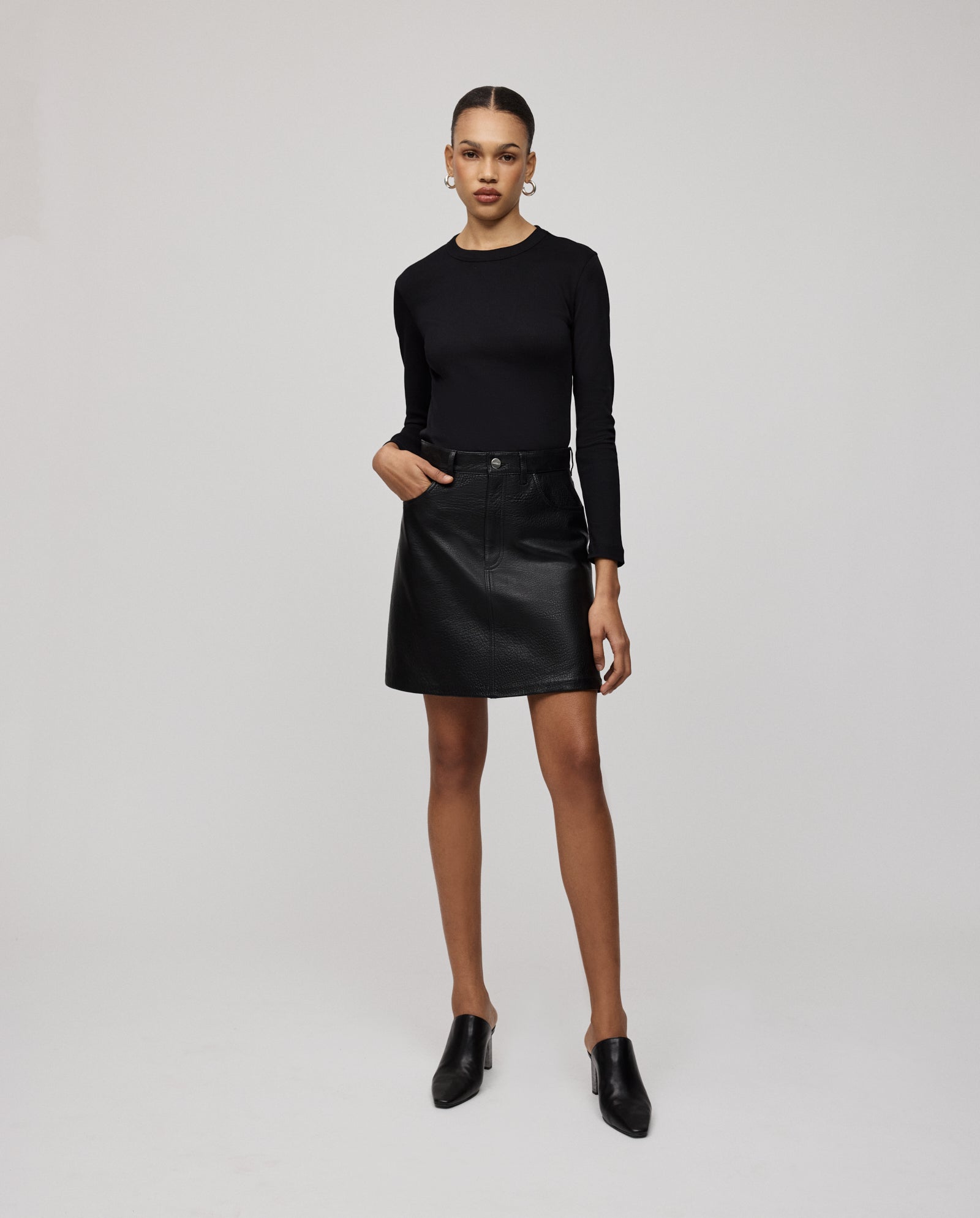 A woman stands against a plain background wearing a black long-sleeve top, black heeled shoes, and the IVY OAK LILOU Skirt—a black premium hide mini skirt with an A-line silhouette.
