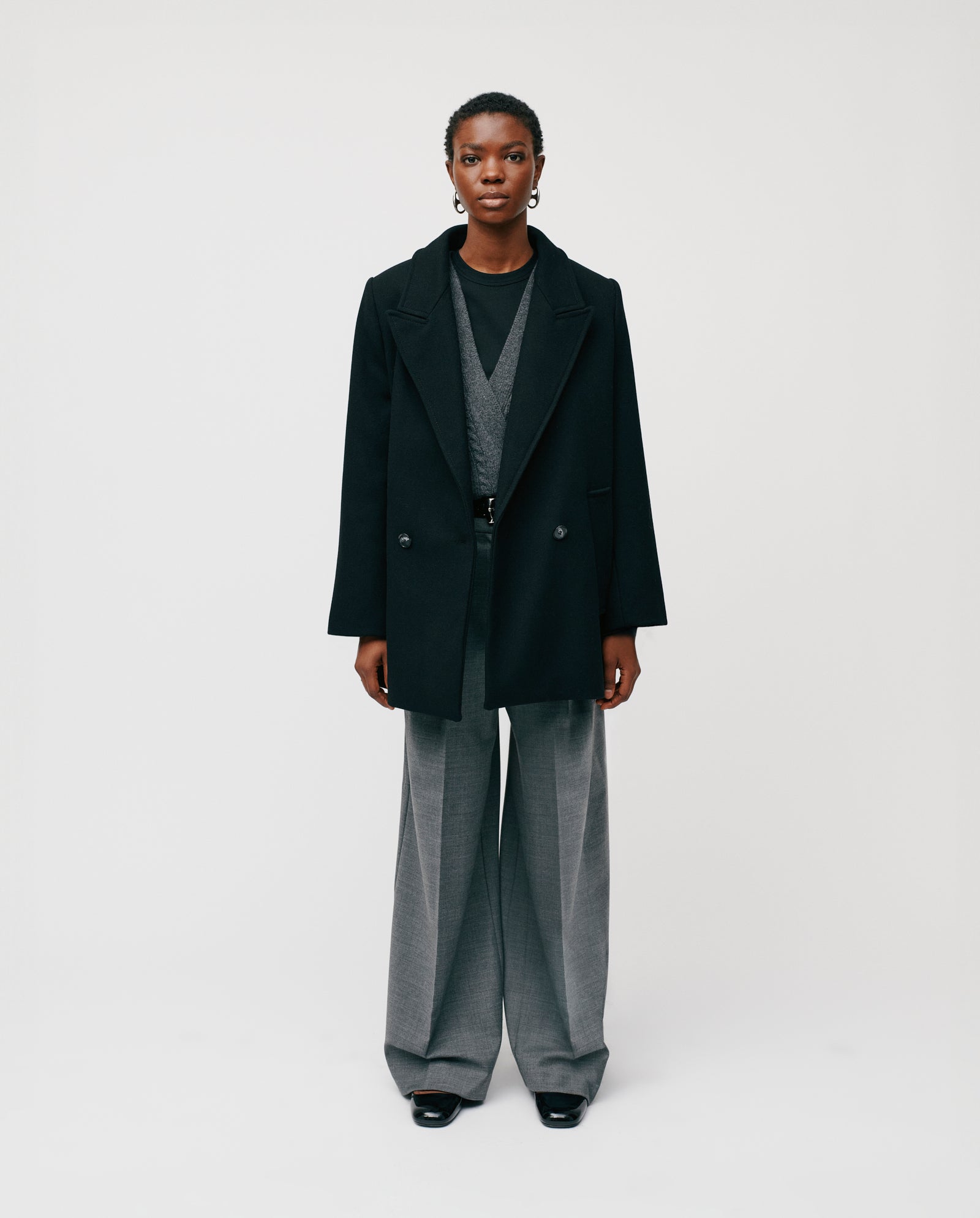 A person stands against a plain background wearing the IVY OAK JENNA black double-breasted jacket, a gray sweater, and wide-leg gray trousers.
