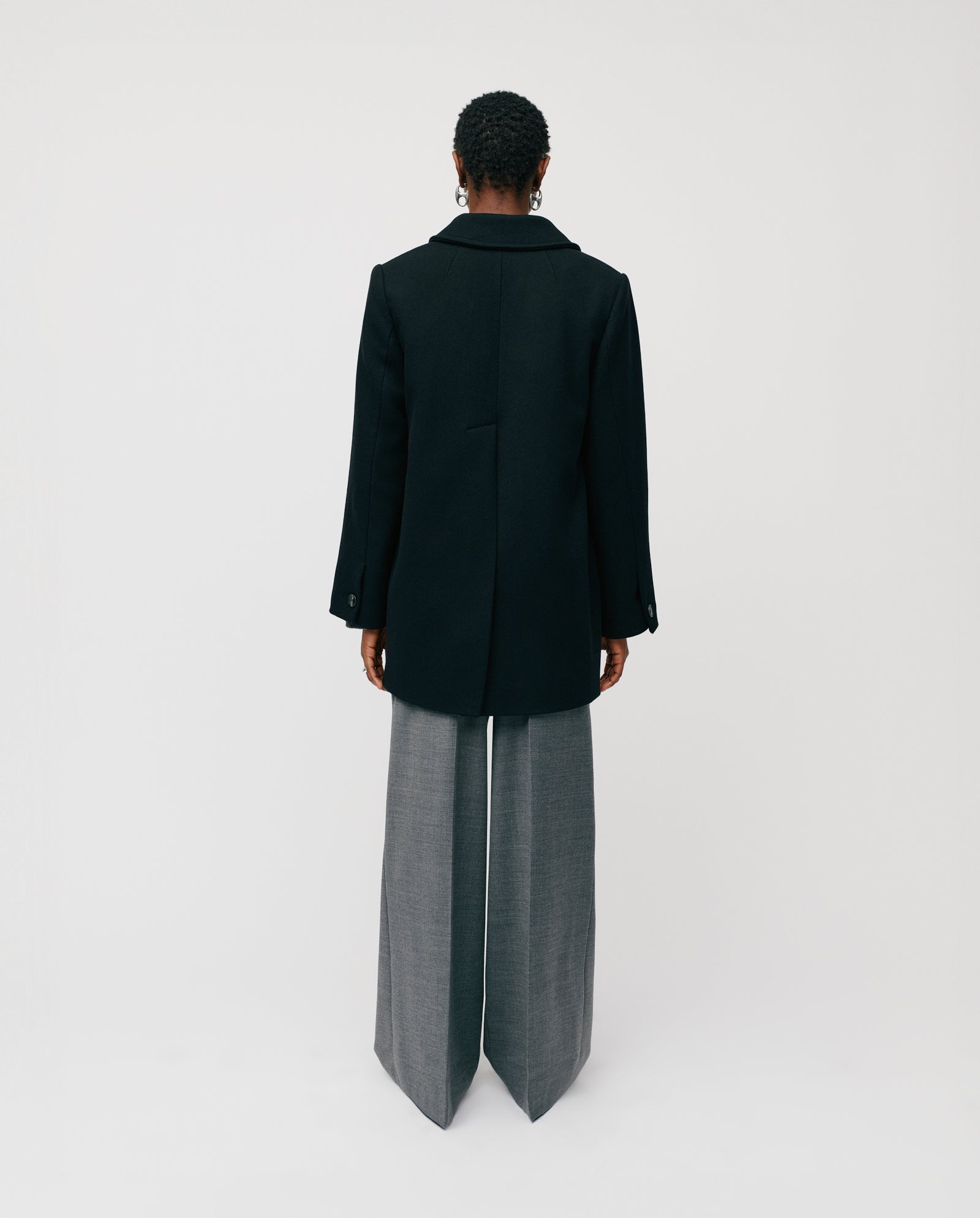 A person stands facing away against a plain white background, wearing the IVY OAK JENNA double-breasted black jacket paired with wide-leg gray pants.