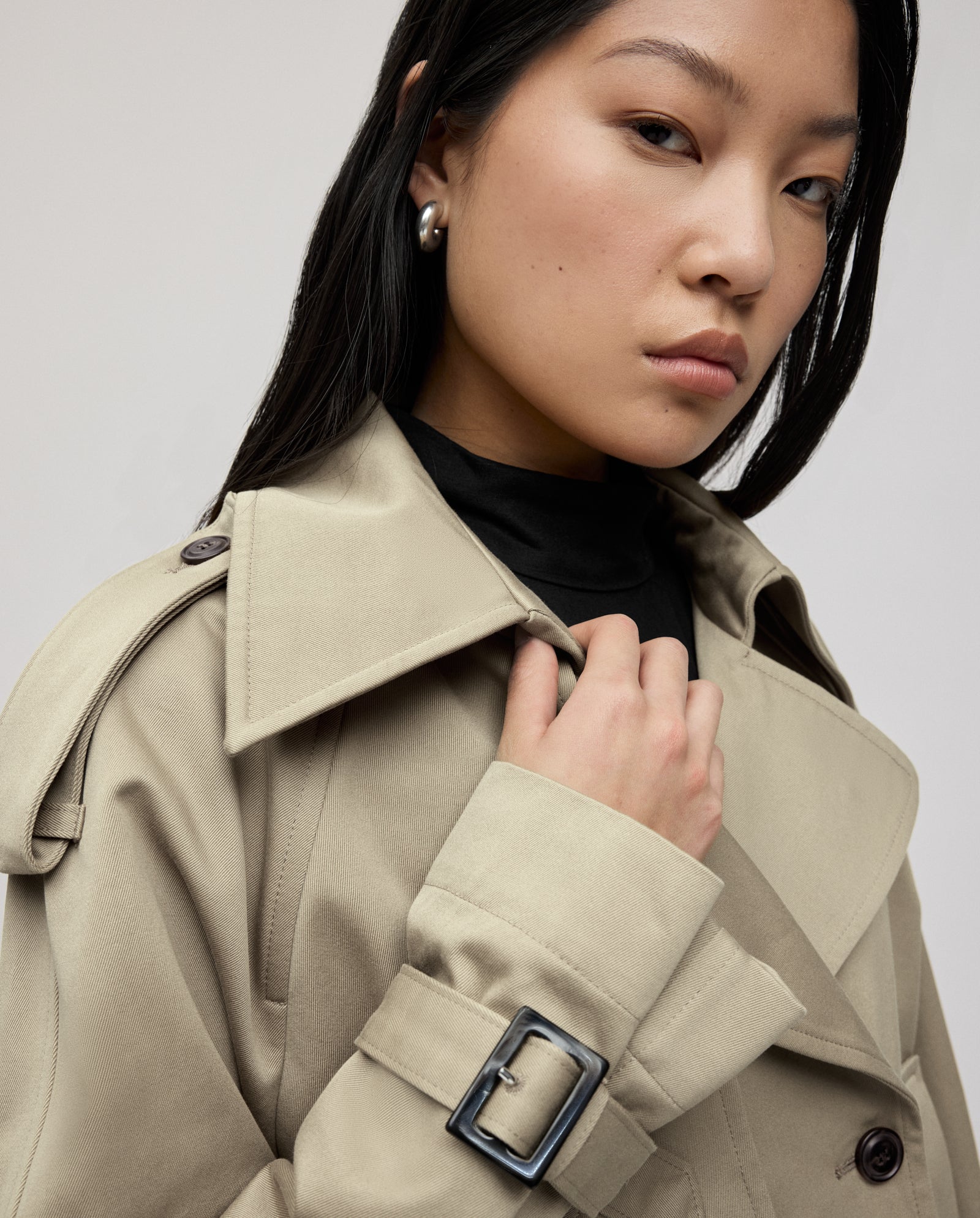 Woman in a beige trench coat and black top, looking confidently at the camera.
