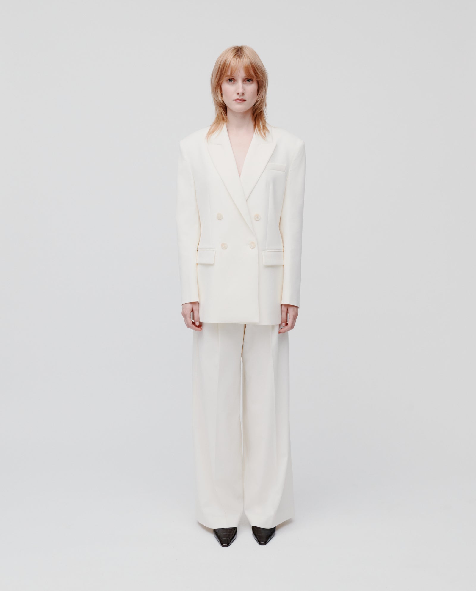 A person wearing the IVY OAK JUDITH Blazer, a white oversized double-breasted blazer in lightweight wool-blend, pairs it with black shoes and stands against a plain white background.